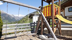 A wooden swing set, a yellow slide, and a playhouse with a mountain backdrop.