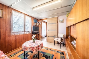 A room with wooden walls, a round table with a floral tablecloth, a TV, a chair, and a cabinet. - Apartment 'Charming T2 With View' with Mountain View, Private Terrace and Wi-Fi (Reignier-Esery)