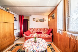 A cozy living room with a floral sofa, a round table with a vase, wooden walls, and a window with lace curtains. - Apartment 'Charming T2 With View' with Mountain View, Private Terrace and Wi-Fi (Reignier-Esery)