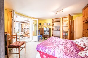 Um quarto com cama, uma cadeira, uma escrivaninha e uma porta aberta que dá para uma cozinha.