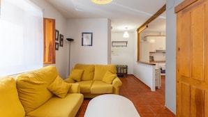 Un salon avec un canapé jaune, un pouf blanc et une cuisine en arrière-plan.