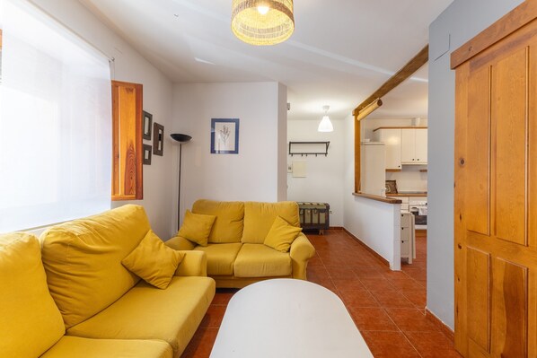 A living room with a yellow sofa, a white ottoman, and a kitchen in the background.