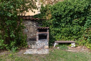 A stone-built outdoor structure with a chimney, surrounded by greenery and a stone wall.