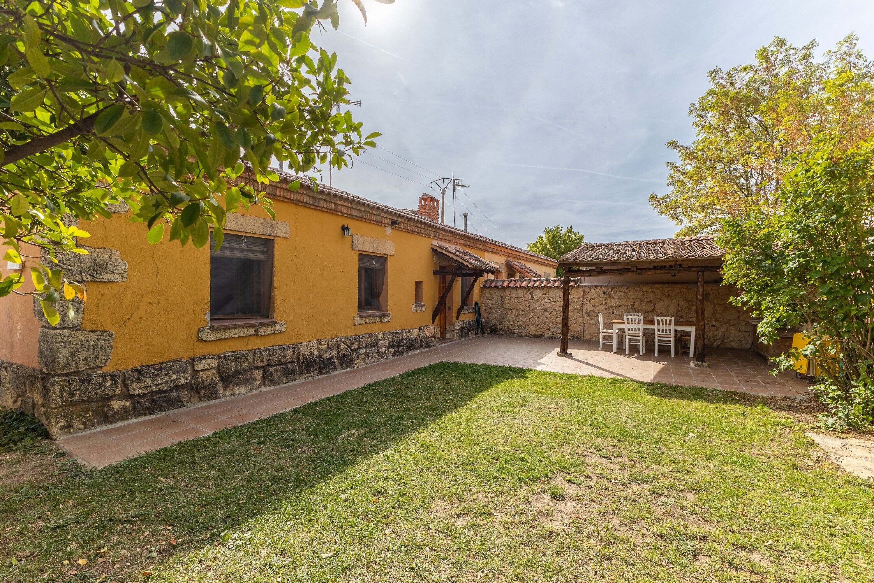 A courtyard with a yellow building, stone walls, a grassy area, and a covered seating area.