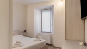 A bedroom with a bed, a bedside table with a lamp, a window, a wall-mounted TV, and a bedside table with a remote control.