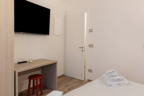 Una habitación de hotel moderna con una cama, un escritorio con un taburete rojo, un televisor montado en la pared y una puerta que da a otra habitación.