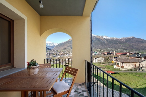 Ein Balkon mit Holztisch und -stühlen, einem Blumentopf und Blick auf eine Stadt und Berge.