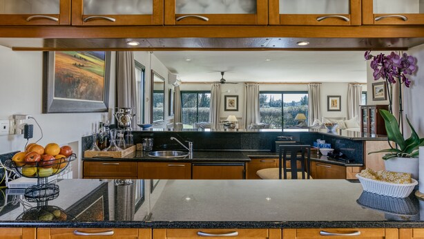 A kitchen with wooden cabinets, a granite countertop, a fruit bowl, and a view of the landscape through large windows. - Villa 'Luxury Golf Villa Torremirona' with Mountain View, Wi-Fi and Air Conditioning (Navata)