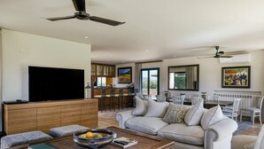A modern living room with a sofa, coffee table, and TV. There is a kitchen area with bar stools and a dining table. - Villa 'Luxury Golf Villa Torremirona' with Mountain View, Wi-Fi and Air Conditioning (Navata)