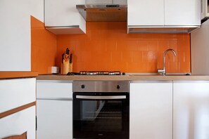 Una cocina con gabinetes blancos, un salpicadero naranja, una campana de acero inoxidable, una estufa a gas, un lavavajillas y un horno empotrado.