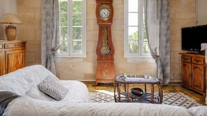 A room with a bed, a wooden cabinet, a table with a lamp, a large wall clock, and a television.