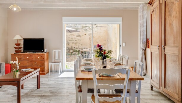 A dining area with a wooden table set for four, white chairs, a television on a wooden cabinet, and a view through a glass door to an outdoor patio.