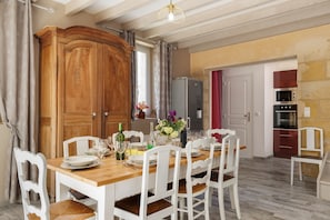 Un coin repas comprenant une table en bois, des chaises blanches, un buffet et un espace cuisine équipé d’appareils électroménagers.