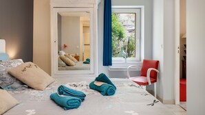 A hotel room with a bed, a mirror, a window with a view, a red chair, and a door leading to another room.