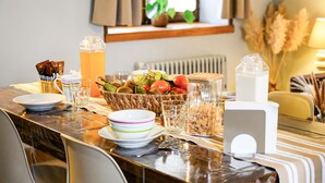 Une table à manger dressée avec des assiettes, des bols, un panier de fruits, un verre d’eau et une carafe de jus.