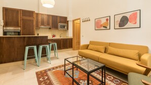 A modern kitchen with wooden cabinets, a microwave, and a sink. There is a yellow sofa and a glass coffee table in the living area.