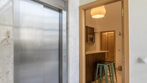A hallway with a metallic elevator, a wooden door leading to a room with a kitchen area, and a stool.