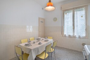 Un coin repas avec une table dressée pour quatre personnes, des chaises jaunes, une nappe blanche et une fenêtre ornée de rideaux à motifs floraux.