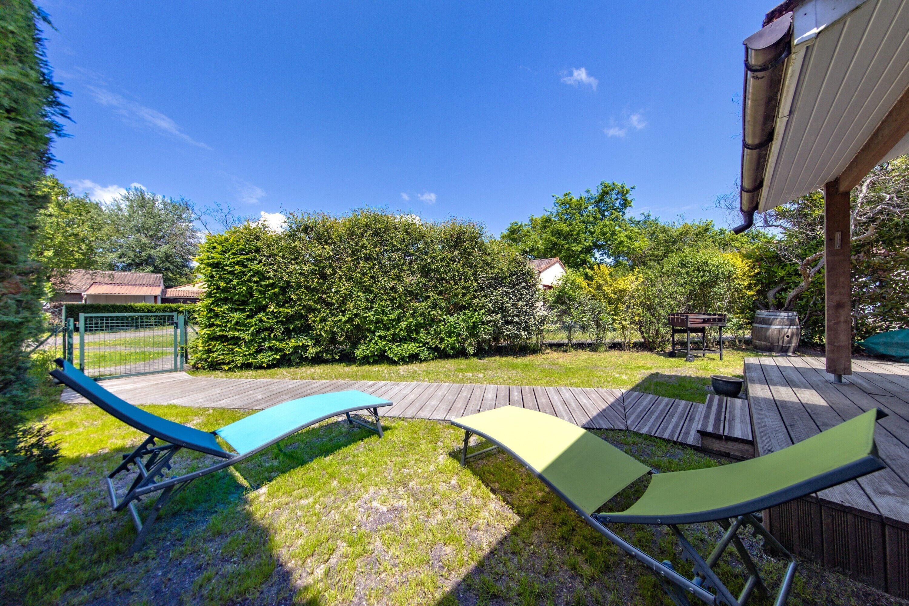 A backyard with a wooden deck, two lounge chairs, a wooden table, and a metal gate.