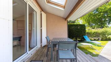 Een overdekt terras met een tafel en stoelen, een schuifpui naar een kamer en een uitzicht op een tuin met een bankje.