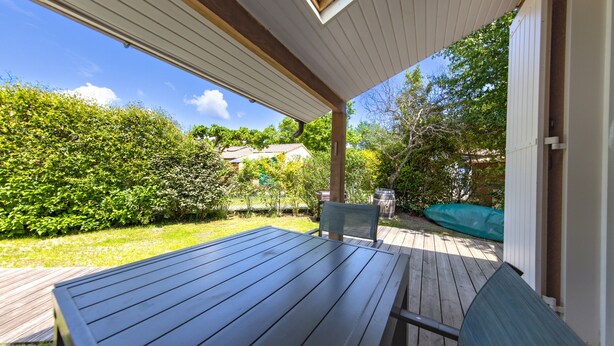 A wooden deck with a table and chairs, surrounded by greenery and a clear sky.
