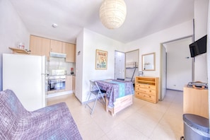 A compact living space with a kitchenette, a dining table, a sofa, a TV, and a trash bin. - Comfortable Chalet Near the Beach and Marina – Garden, Parking, Leisure Facilities (Hourtin)