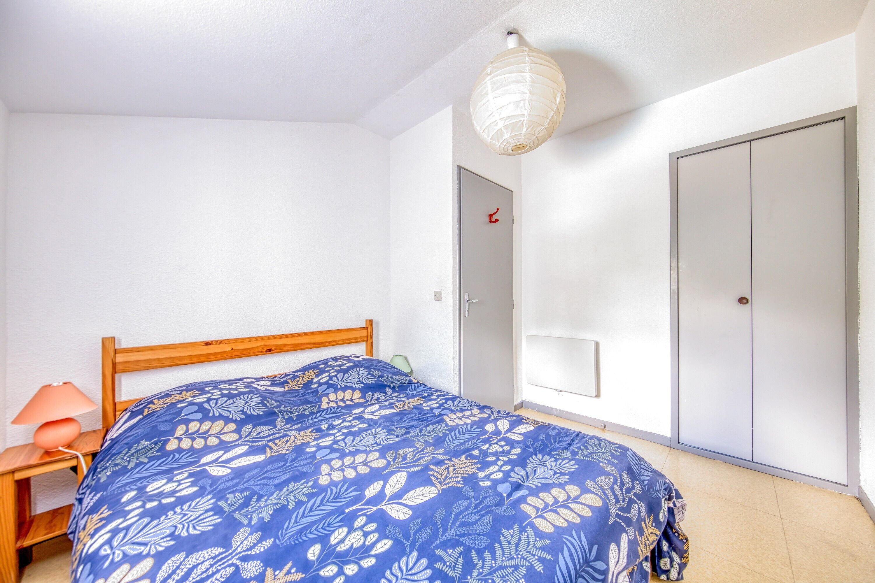 A bedroom with a bed, a nightstand, a lamp, a wardrobe, and a door.