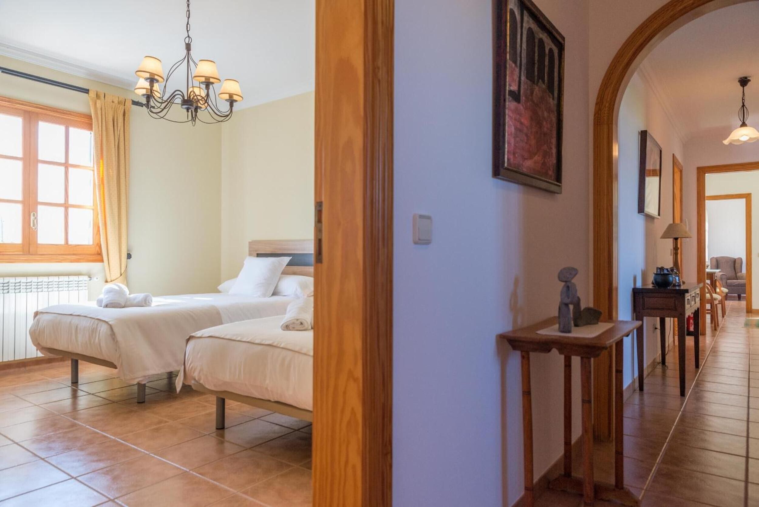 Una habitación de hotel con dos camas, una araña, un cuadro y un pasillo con una puerta de madera.