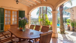 A covered patio with a wooden table and chairs, a window with green shutters, and potted plants. - Villa 'Vernissa 10p' with Mountain View (Santa Margalida)