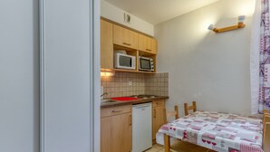 A compact kitchen with wooden cabinets, a microwave, a small refrigerator, and a dining table with a checkered tablecloth. - Apartment 'A1 Renard' with Mountain View, Shared Garden and Wi-Fi (Le Bourg-d'Oisans)