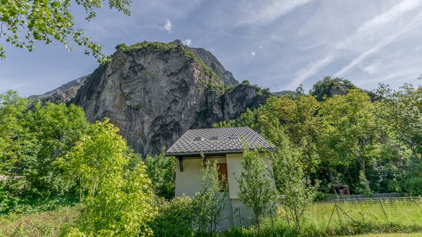 A small building with a sloped roof, surrounded by lush greenery and a prominent mountain in the background. - Apartment 'B3 Chamois' with Mountain View, Shared Garden and Wi-Fi (Le Bourg-d'Oisans)