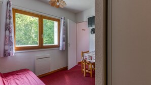 A bedroom with a bed, a window with curtains, a radiator, a wooden table, and a door. - Apartment 'B3 Chamois' with Mountain View, Shared Garden and Wi-Fi (Le Bourg-d'Oisans)