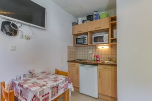 A compact kitchen with a microwave, oven, and a small refrigerator. There is a table with a quilted tablecloth and a chair. - Apartment 'B3 Chamois' with Mountain View, Shared Garden and Wi-Fi (Le Bourg-d'Oisans)