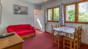 Een kamer met een rode bank, een eettafel met stoelen, een plafondlamp, een schilderij aan de muur en een raam met gordijnen.