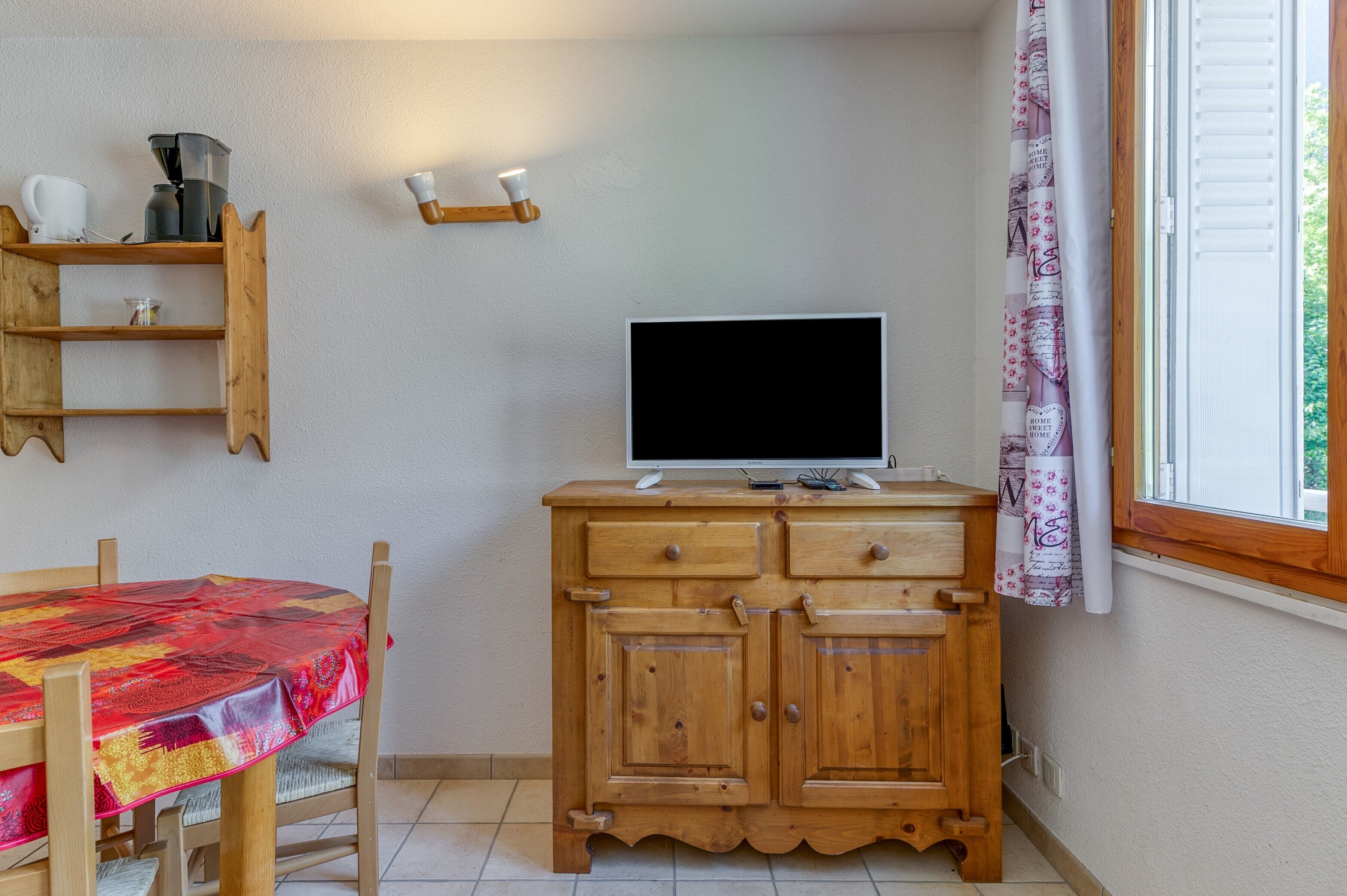A room with a wooden cabinet, a television on top, a table with a red tablecloth, and a window with a curtain.