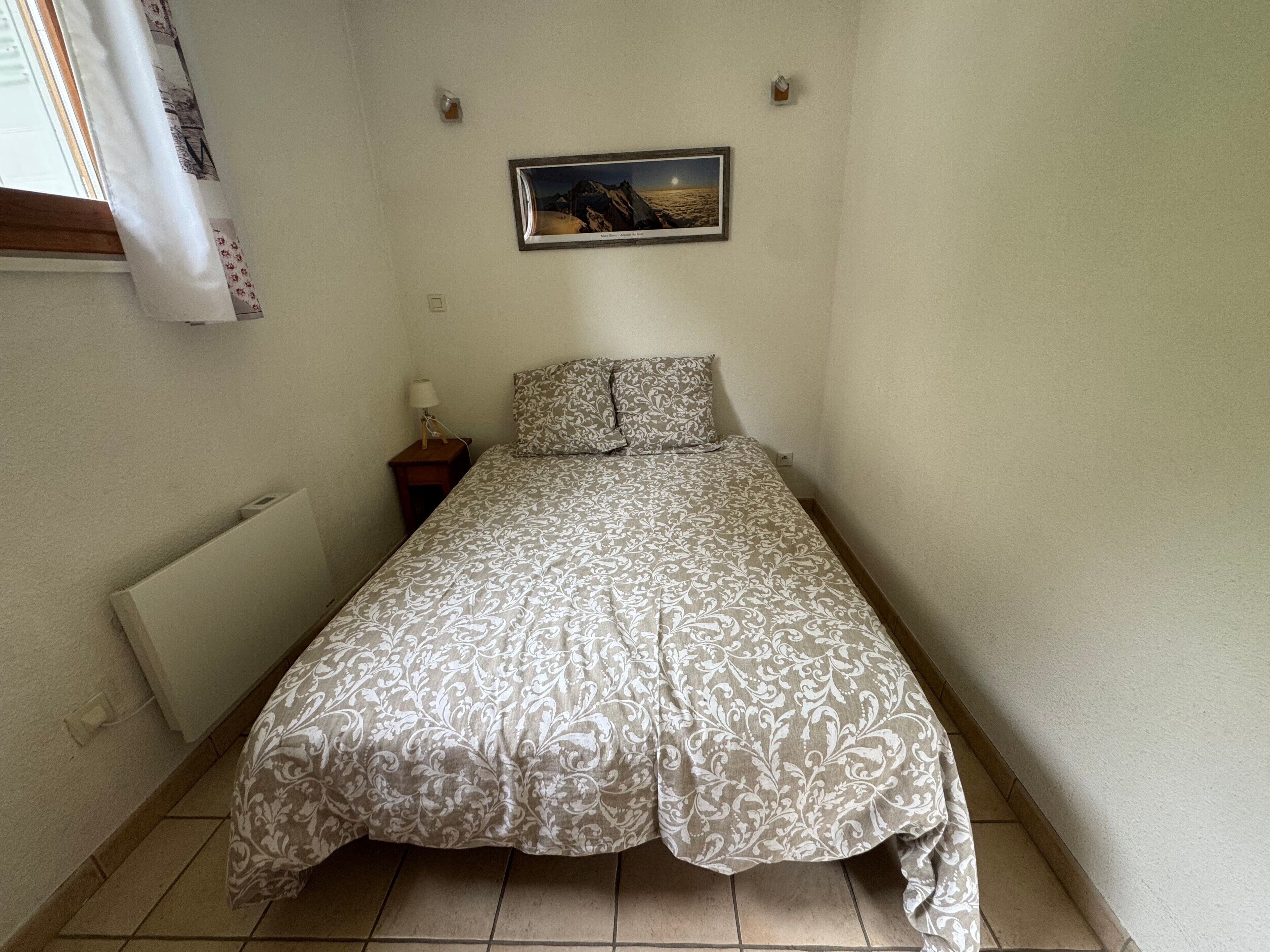 A small, narrow room with a bed, a nightstand, a lamp, a picture on the wall, and a radiator.