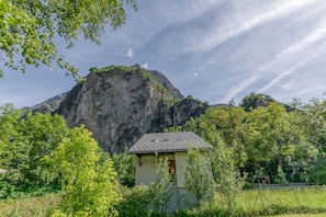 A small building with a sloped roof, surrounded by lush greenery and a prominent mountain in the background.