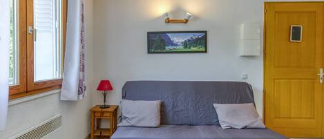 A room with a bed, a small table with a lamp, a picture on the wall, a door, and a window with curtains.
