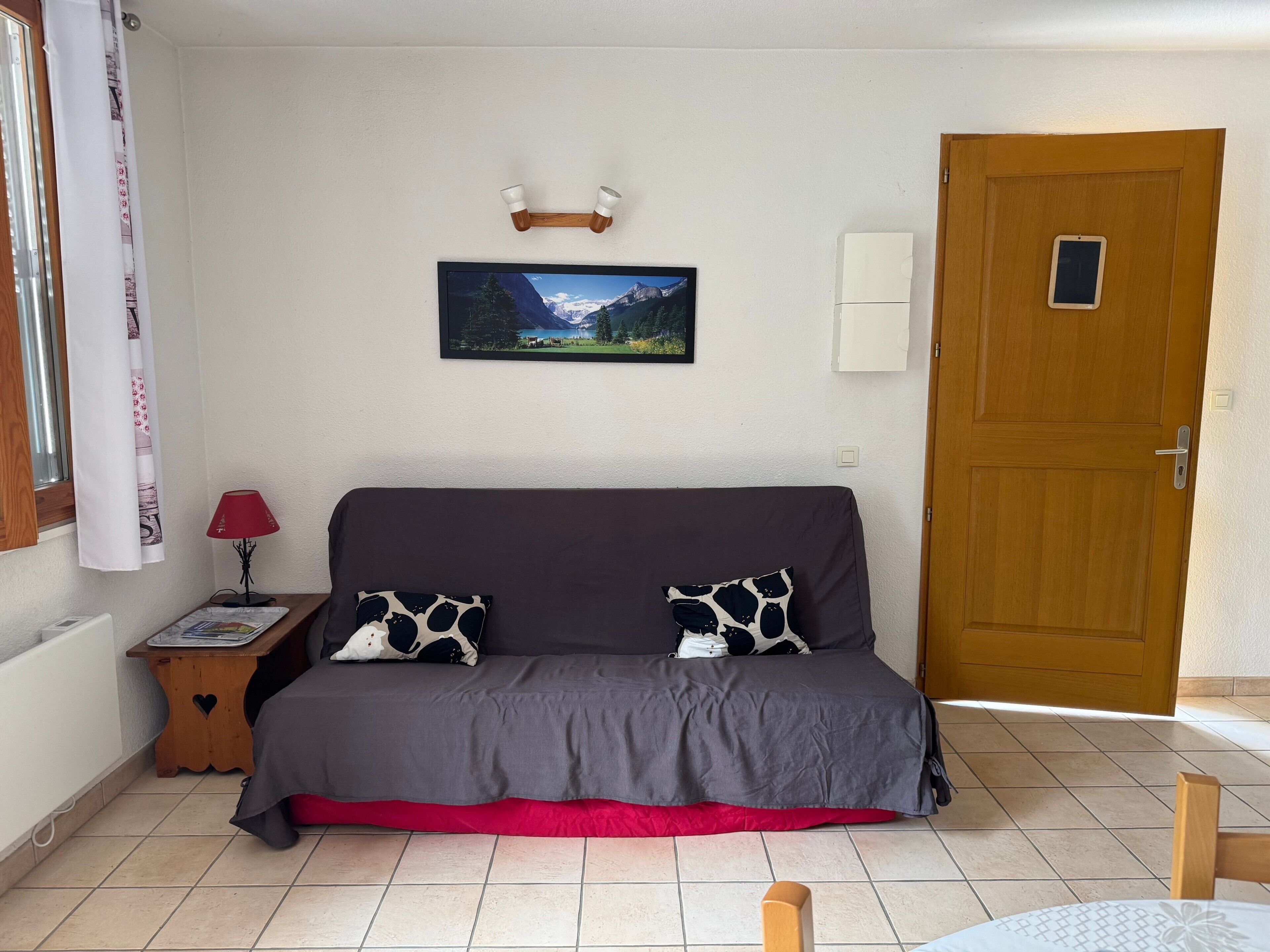 A room with a grey sofa, a red cushion, a wooden side table with a lamp, a framed picture on the wall, and a wooden door.