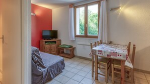 Ein Zimmer mit einem Bett, einem Fernseher auf einer Holzkommode, einem Esstisch mit Stühlen und einem Fenster mit Vorhängen.