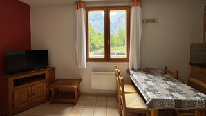 Ein Zimmer mit Fernseher auf einer Holzkommode, einem kleinen Holzhocker, einem Tisch mit gemustertem Tischtuch und einem Fenster mit Vorhängen.