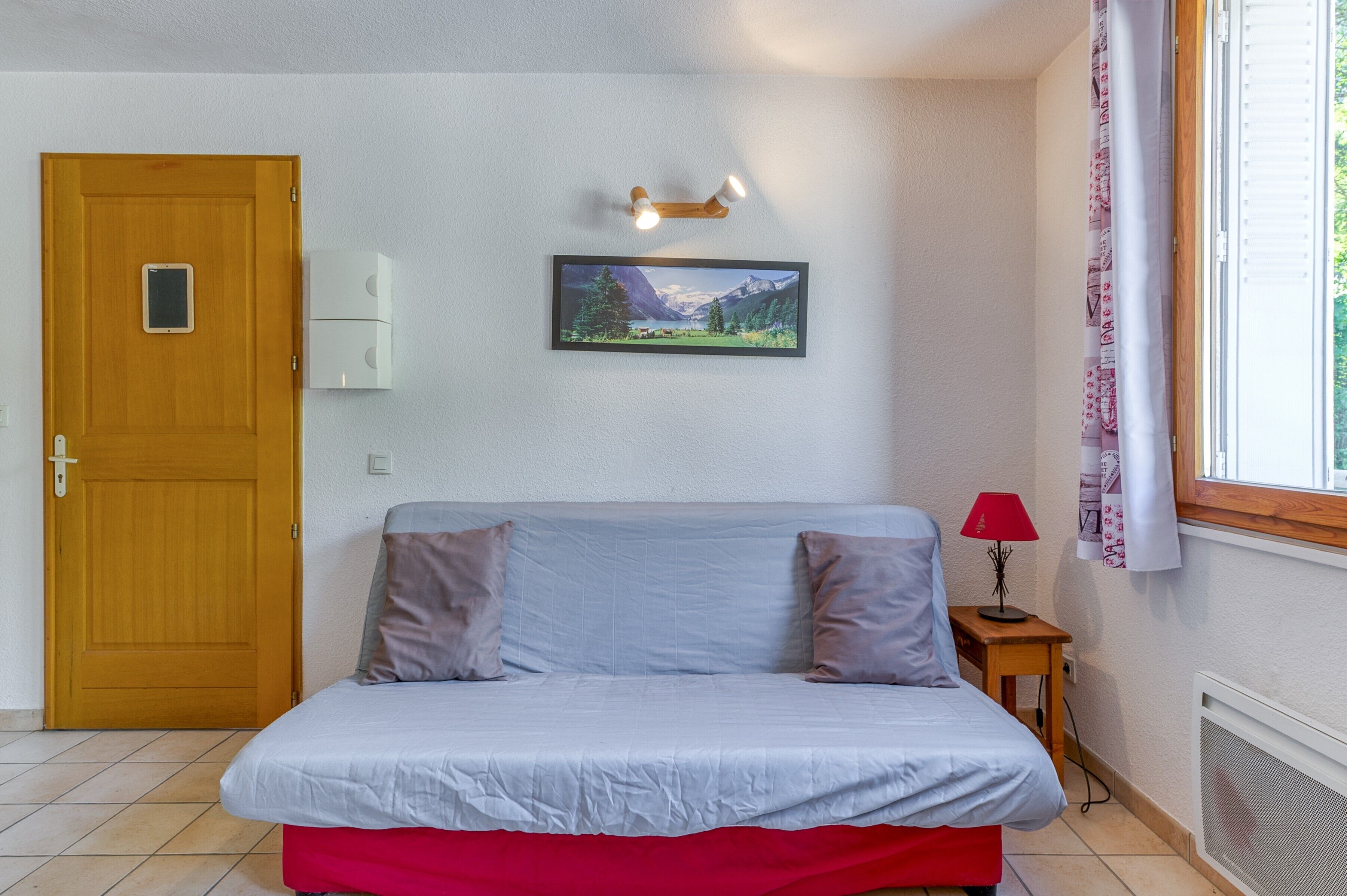 A room with a bed, a door, a wall-mounted light fixture, a picture, a window with curtains, and a small table with a lamp.