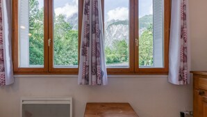 A room with a window, curtains, a radiator, and a small wooden table. - Apartment 'B4 Ecureuil' with Mountain View, Shared Garden and Wi-Fi (Le Bourg-d'Oisans)