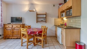 A compact kitchen with wooden cabinets, a dining table with chairs, a microwave, and a television. - Apartment 'B4 Ecureuil' with Mountain View, Shared Garden and Wi-Fi (Le Bourg-d'Oisans)