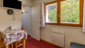 A room with a table covered in a patterned tablecloth, a window with a view of trees, a radiator, and a television mounted on the wall.