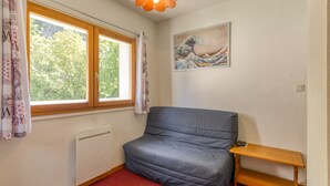 A room with a grey sofa, a wooden shelf, a radiator, a window with curtains, and a painting on the wall.