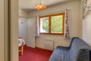 Une pièce comprenant un lit, une table recouverte d’une nappe, un radiateur, une fenêtre avec des rideaux et un luminaire suspendu.