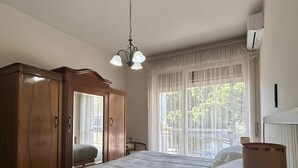 Une chambre à coucher comprenant un lit, une armoire en bois, une table de chevet avec une lampe, une fenêtre avec des rideaux légers et un appareil de climatisation.