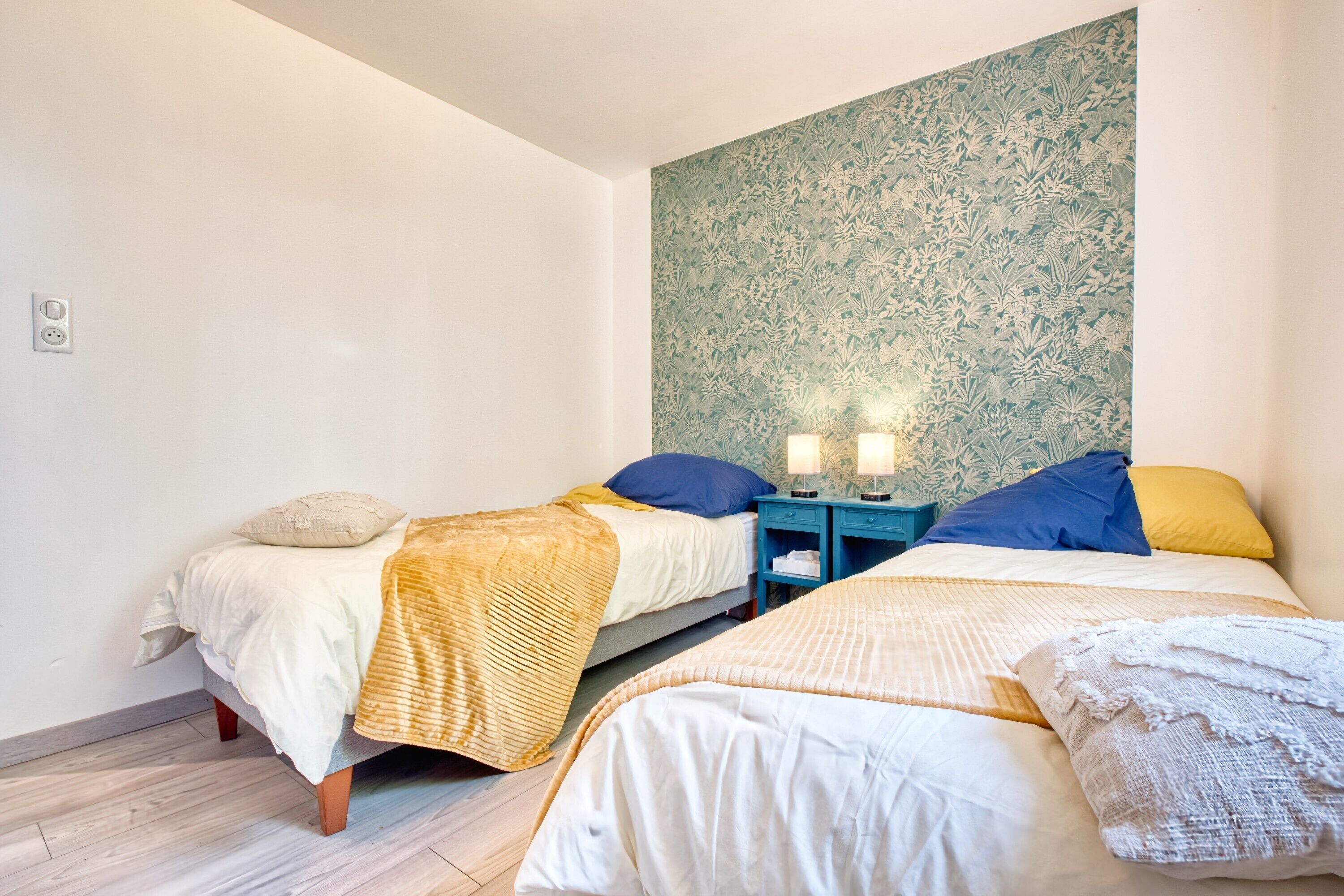 A room with two beds, a patterned wall, and a nightstand with lamps.