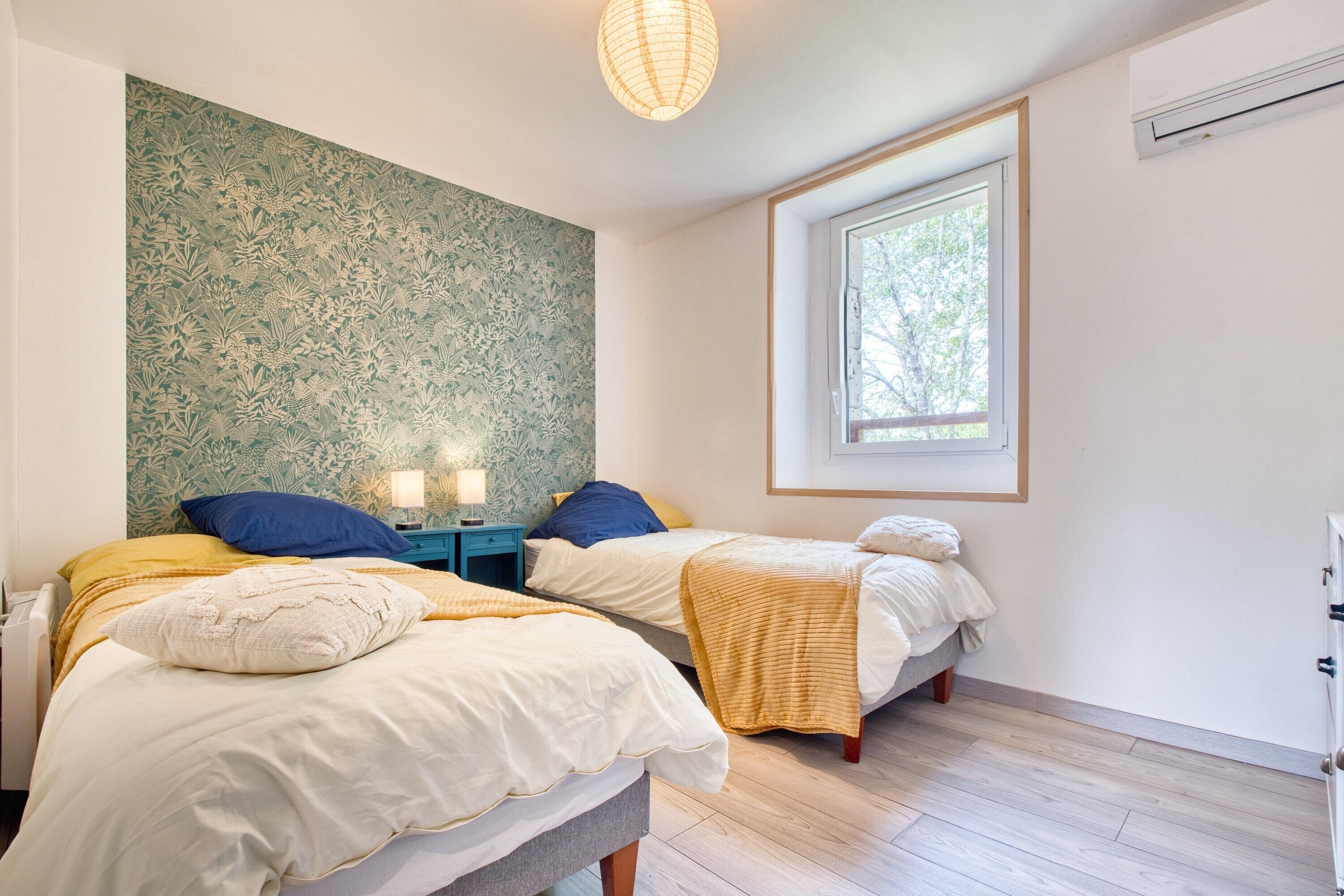 Slaapkamer met twee bedden, een raam, een wand met bloemenprint en een plafondlamp.